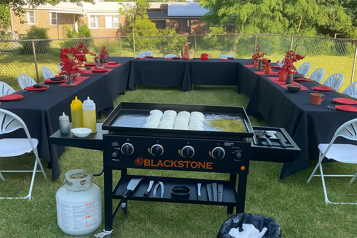 Outdoor dining area setup on the lawn. Long tables covered with white tablecloths, paired with black chairs, showing a casual gathering scene